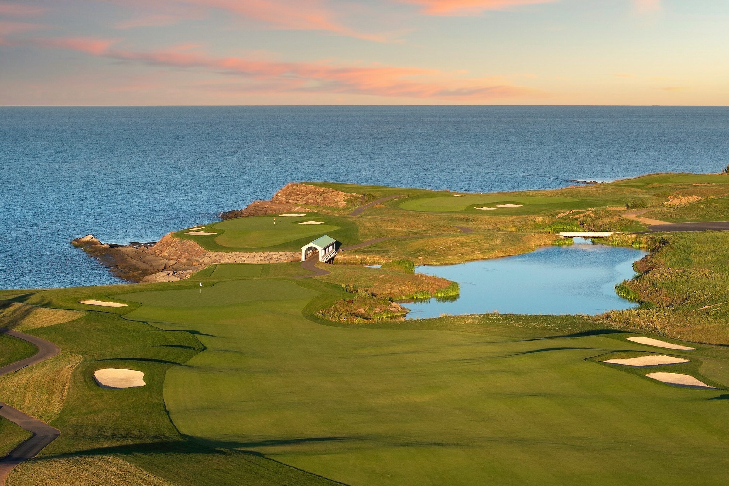Aerial view of Ocean Course greens on the rocky edge of the Northumberland Strait, with a pond below them, in low golden light