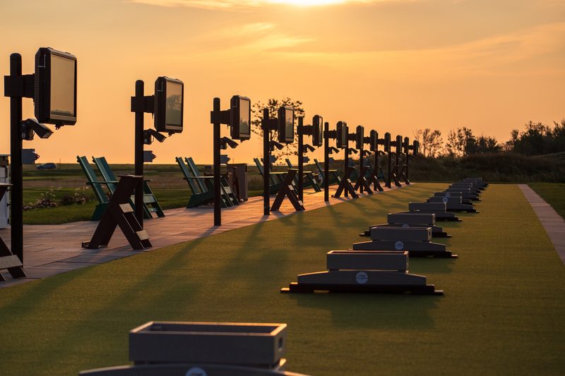 TrackMan screens on posts at each hitting bay on the Fox Harb'r driving range at sunset, with Adirondack chairs behind